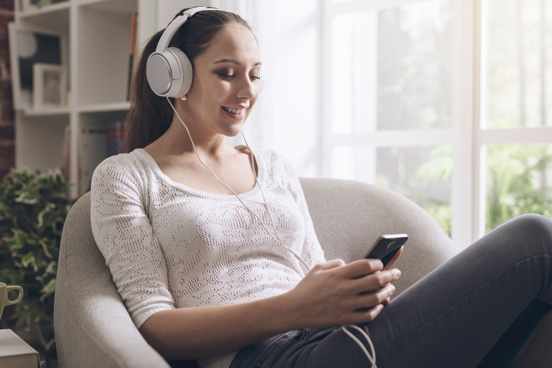 Happy young woman listening to music at home, she is connecting with her smartphone and wearing headphones